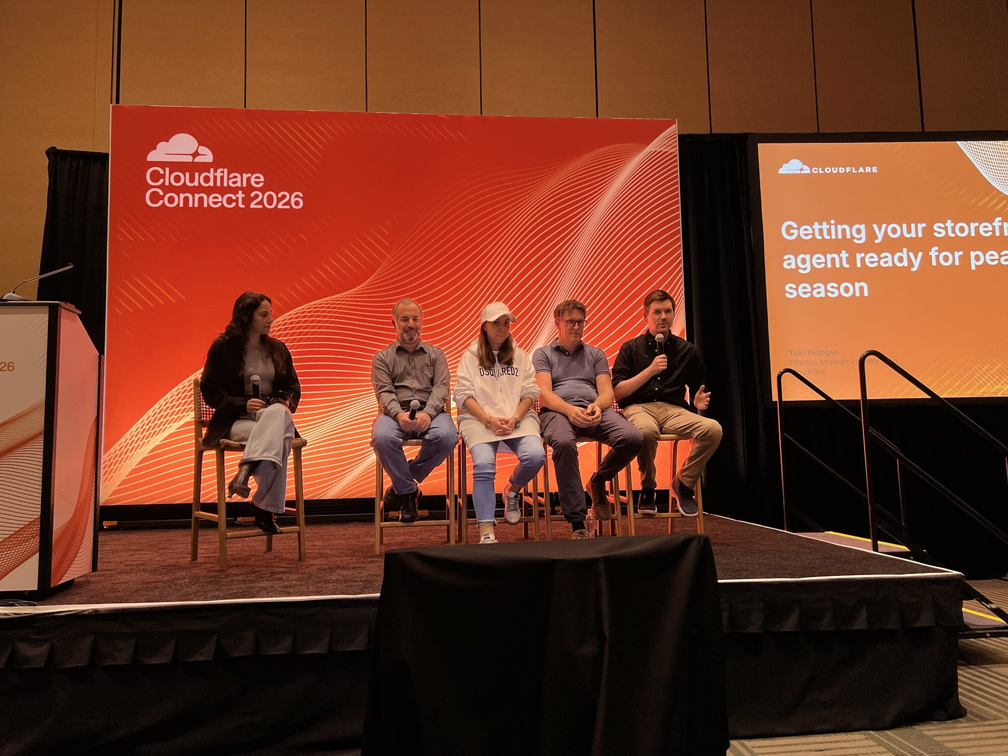 Five panelists seated on stools on the Cloudflare Connect 2026 stage in front of an orange backdrop, holding microphones, with the session title on a screen to the right.